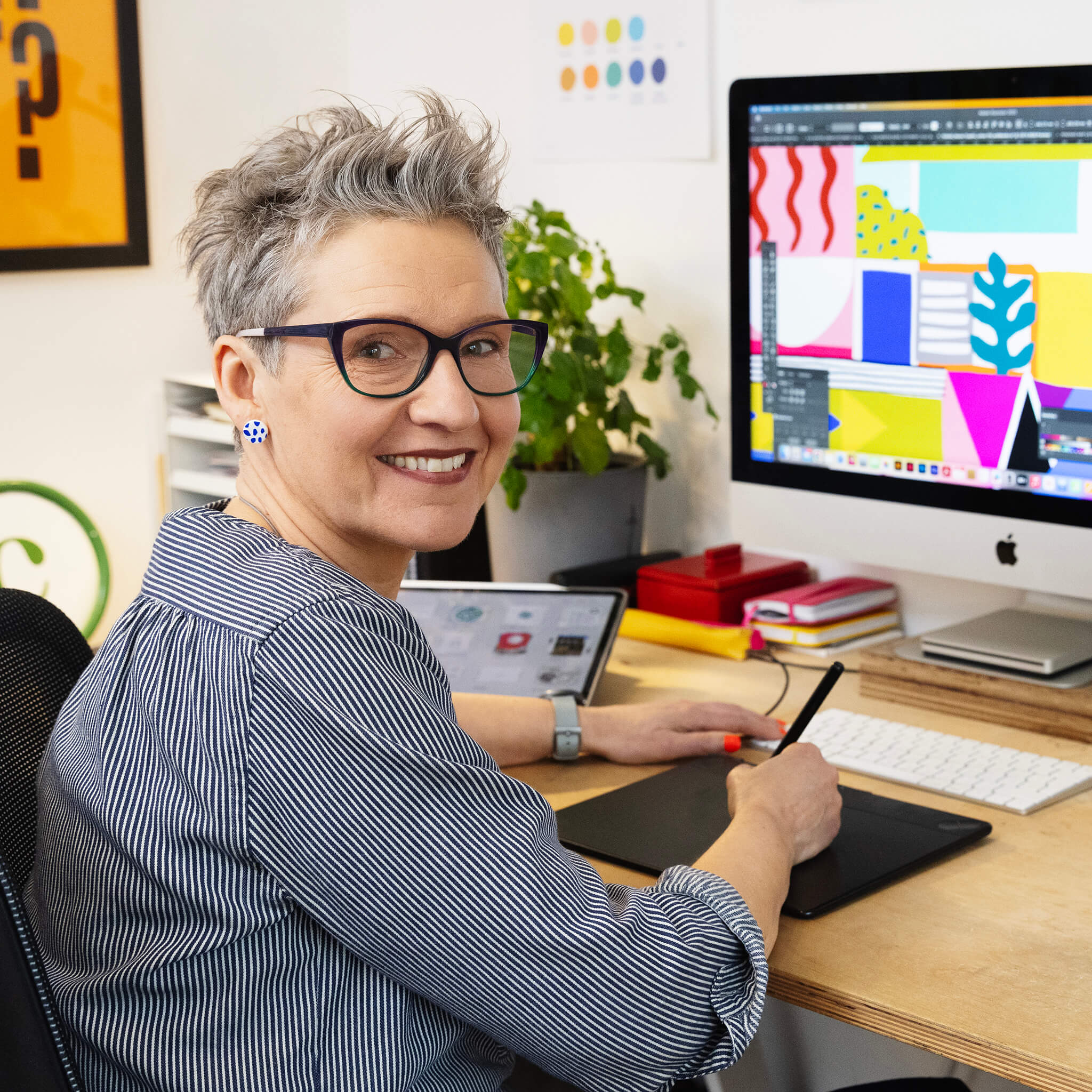 Photo of digital illustrator Carys Tait sitting working at desk. Colourful graphic illustration on the computer screen